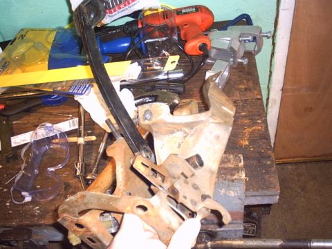 1991 Ford LTD Crown Victoria - A removed brake and clutch pedal assembly with its stamped-steel mounting bracket and curved pedal arm sits on a cluttered wooden workbench…