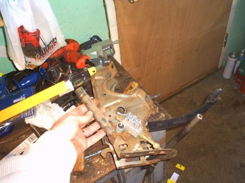 1991 Ford LTD Crown Victoria - A rusty stamped-steel pedal support bracket clamped in a bench vise on a wooden workbench while a hand steadies it during disassembly, with…