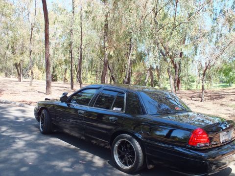 2003 Ford Crown Victoria - Blacked-out full-size sedan shown from the rear quarter on a wooded park road.