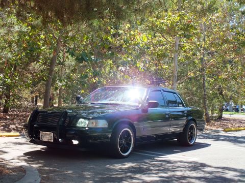 2003 Ford Crown Victoria - Lowered black full-size sedan with push bar parked among sunlit trees.