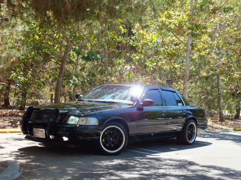 2003 Ford Crown Victoria - Front-quarter view of a lowered black sedan with push bar and spotlight under trees.