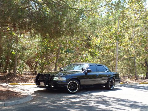 2003 Ford Crown Victoria - Blacked-out Crown Victoria sedan with push bar and dark wheels sits on tree-shaded pavement