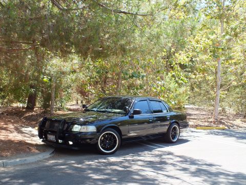 2003 Ford Crown Victoria - Dark sedan with push bar and black wheels rests on shaded asphalt near woods