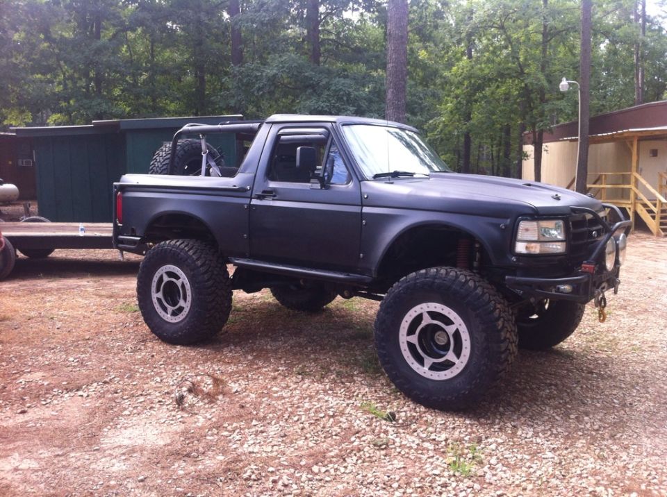Modified black Ford Bronco with large off-road tires and a roll cage, parked on gravel