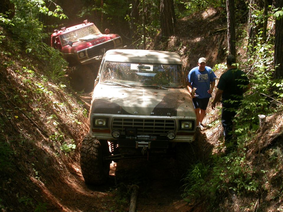 1979 Ford F-150 with camouflage paint, lifted suspension, and winch, driving on a muddy trail with another red truck behind it