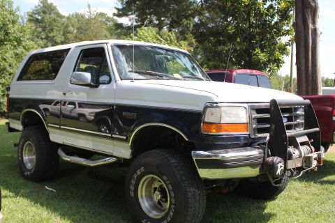 1994 Ford Bronco - A two-tone Ford Bronco is parked on a grassy area with trees in the background.