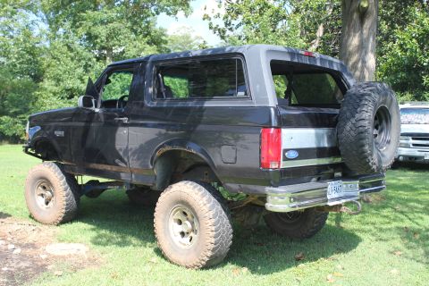 1994 Ford Bronco - A lifted black Ford Bronco is parked on a grassy area with trees in the background.
