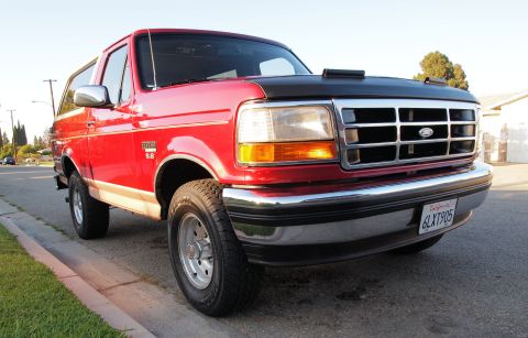 1994 Ford Bronco - This red Ford Bronco is parked curbside on a sunny day, showing its front quarter panel and grille.