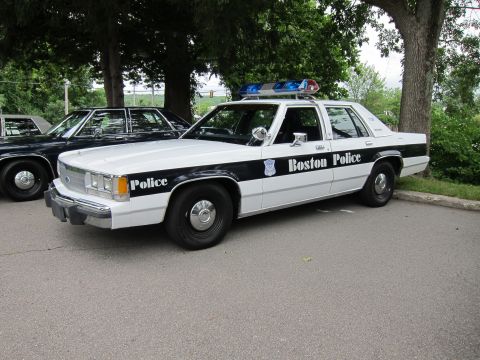 1991 Ford LTD Crown Victoria - black-and-white Ford LTD Crown Victoria marked Boston Police, with roof lightbar and spotlight, parked under trees.