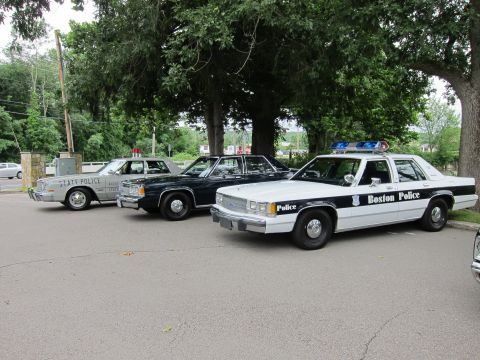 1991 Ford LTD Crown Victoria - black-and-white Boston Police Ford sedan parked alongside a State Police cruiser and other vintage police cars under trees.