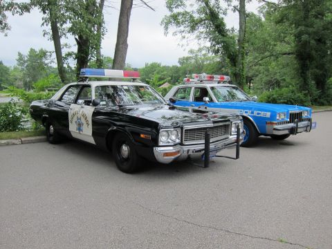 1991 Ford LTD Crown Victoria - black-and-white California Highway Patrol-style sedan parked beside a blue and white POLICE cruiser among trees.