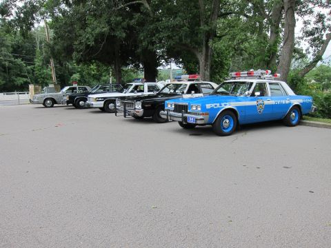 1991 Ford LTD Crown Victoria - row of vintage police cruisers parked on pavement, led by a blue POLICE sedan with a roof lightbar.