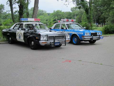 1991 Ford LTD Crown Victoria - black-and-white Highway Patrol sedan with push bumper parked next to a blue and white POLICE cruiser under trees.