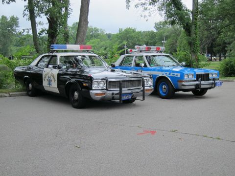 1991 Ford LTD Crown Victoria - black-and-white Highway Patrol sedan with push bumper beside a blue and white POLICE cruiser, parked among green trees.