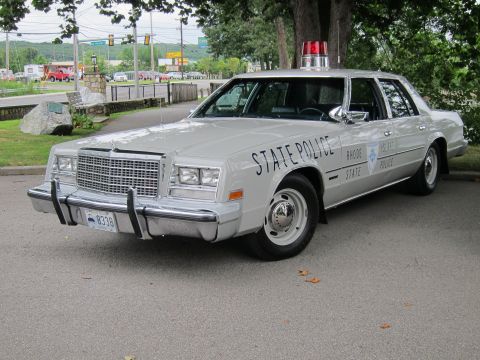 1991 Ford LTD Crown Victoria - silver Chrysler-style sedan marked RHODE ISLAND STATE POLICE with a single red roof light, parked near a roadway.