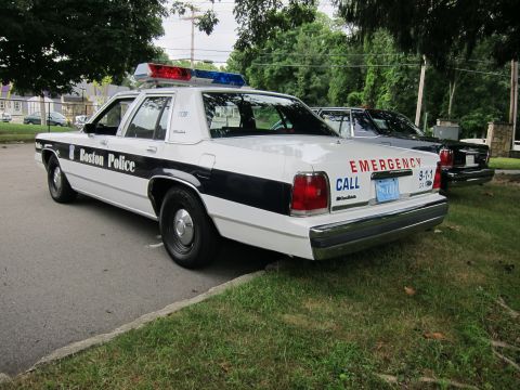 1991 Ford LTD Crown Victoria - Rear three-quarter view of a black-and-white Boston Police Ford sedan with red EMERGENCY CALL 9-1-1 markings on the trunk.