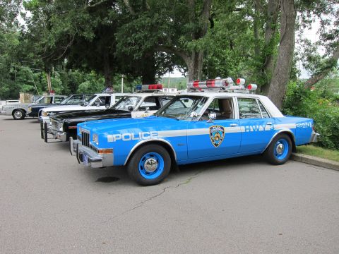 1991 Ford LTD Crown Victoria - restored blue-and-white NYPD Highway Patrol sedan marked 'POLICE HWY 3' parked beside other vintage police cars on a tree-lined lot.