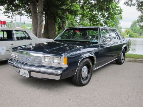 1991 Ford LTD Crown Victoria - Front quarter view of a dark blue Ford LTD Crown Victoria sedan parked next to a marked 'Island Police' cruiser on a lakeside lot.