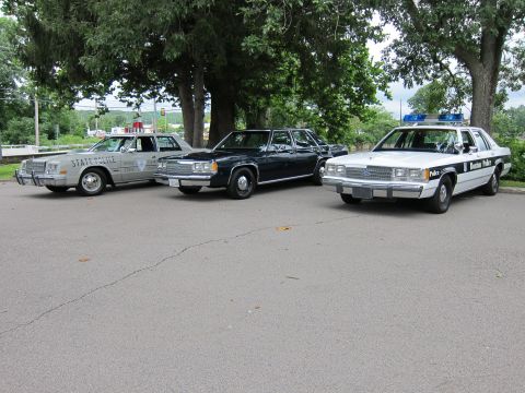1991 Ford LTD Crown Victoria - Three vintage cruisers parked side by side on pavement: a silver State Police sedan, a dark unmarked sedan, and a black-and-white Boston Po…