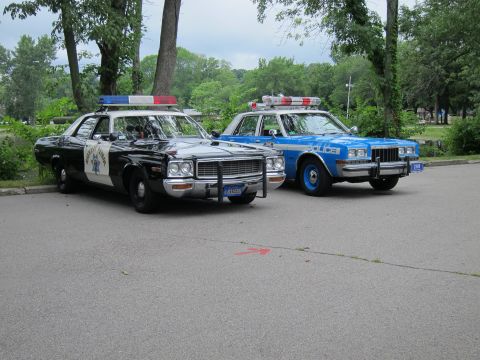 1991 Ford LTD Crown Victoria - black-and-white Highway Patrol sedan parked next to a blue NYPD 'POLICE' cruiser, both with roof lightbars, on a park lot under trees.