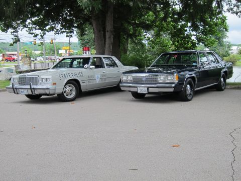 1991 Ford LTD Crown Victoria - silver 'Rhode Island State Police' sedan with single roof light parked beside a black Ford Crown Victoria sedan on a lakeside lot.