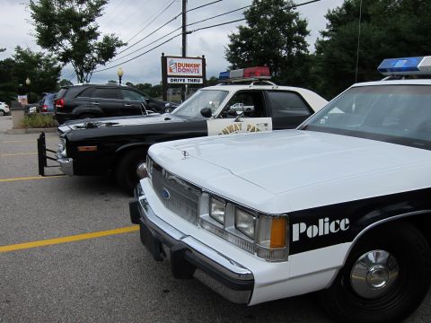 1991 Ford LTD Crown Victoria - Close front view of a white Ford 'Police' cruiser with roof lights parked beside a black police car at a lot with a Dunkin' Donuts sign.