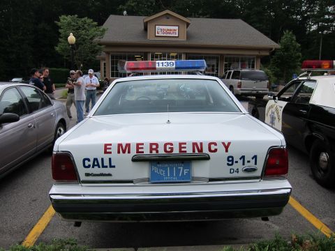 1991 Ford LTD Crown Victoria - Rear view of a white Ford police sedan lettered 'EMERGENCY CALL 9-1-1' with a Massachusetts official plate, parked at a Dunkin' Donuts with…