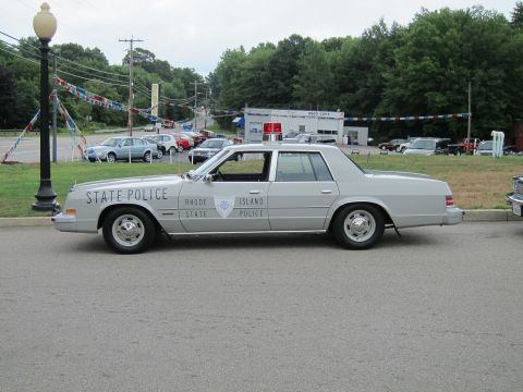 1991 Ford LTD Crown Victoria - Full side profile of a silver 'Rhode Island State Police' four-door sedan with a single roof light, parked near a used-car lot with banners.