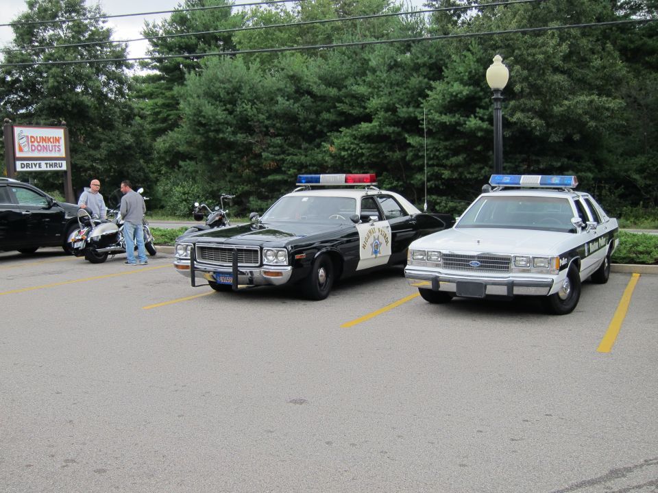 Two vintage police cars, a black highway patrol and a white cruiser, at a Dunkin Donuts lot