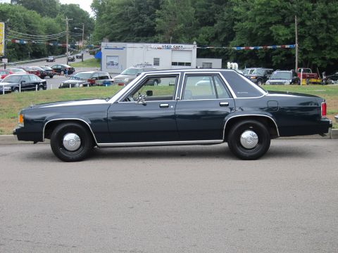 1991 Ford LTD Crown Victoria - Driver-side profile of a dark blue Ford LTD Crown Victoria sedan with dog-dish hubcaps parked on pavement near a used-car dealership.