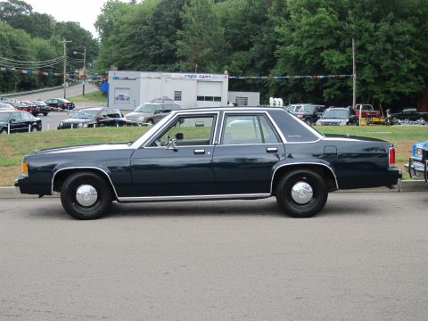 1991 Ford LTD Crown Victoria - Side view of a dark blue Ford LTD Crown Victoria four-door sedan parked along a road in front of a used-car lot with strung banners.