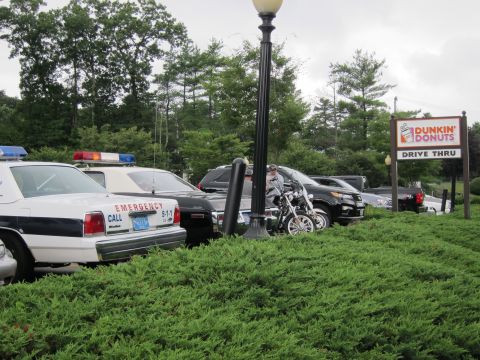 1991 Ford LTD Crown Victoria - White police sedans lettered 'EMERGENCY CALL 911' and a motorcycle parked behind low shrubs, with a Dunkin' Donuts Drive Thru sign at right.