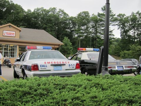 1991 Ford LTD Crown Victoria - white Ford lettered 'EMERGENCY CALL 9-1-1' parked next to a black Highway Patrol cruiser at a Dunkin' Donuts lot behind a hedge.