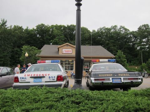 1991 Ford LTD Crown Victoria - white 'EMERGENCY CALL 911' Ford and a black-and-white Highway Patrol sedan parked on either side of a lamppost outside a Dunkin' Donuts.