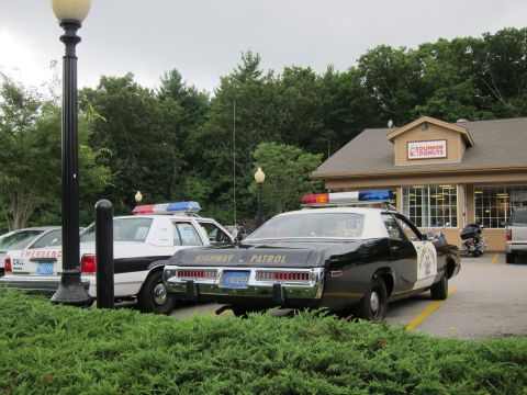 1991 Ford LTD Crown Victoria - Rear quarter view of a black-and-white 'Highway Patrol' cruiser beside a white police Ford at a Dunkin' Donuts lot with a lamppost and hedg…
