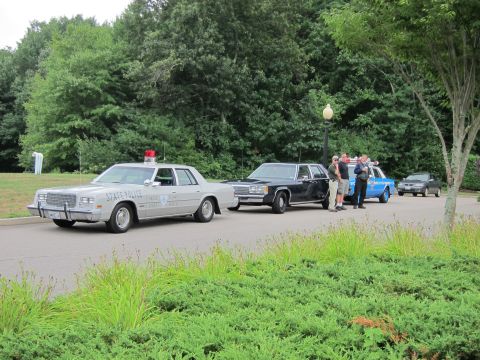 1991 Ford LTD Crown Victoria - silver 'State Police' sedan leads a black sedan and a marked cruiser parked on a road; two men stand between them beside a grassy tree line.