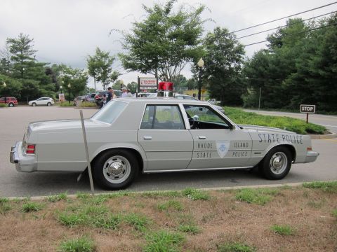 1991 Ford LTD Crown Victoria - Profile of a silver four-door Rhode Island State Police sedan with a red roof light parked on grass near a stop sign.