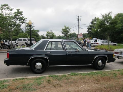 1991 Ford LTD Crown Victoria - Profile of a dark blue four-door Ford police sedan with plain black wheels parked under a cloudy sky.