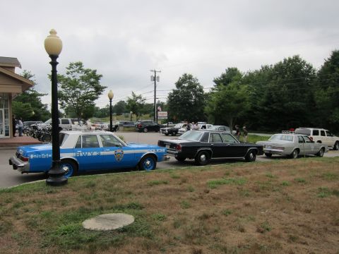 1991 Ford LTD Crown Victoria - blue NYPD cruiser leads a row of parked vintage police cars and motorcycles under an overcast sky.
