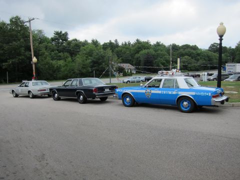 1991 Ford LTD Crown Victoria - blue NYPD cruiser sits at the head of three parked vintage police sedans on a paved lot near trees.