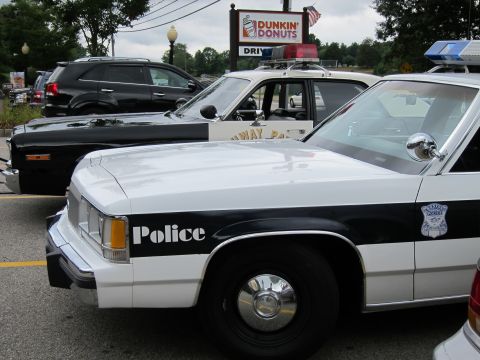 1991 Ford LTD Crown Victoria - Close view of a white-and-black police cruiser fender lettered Police, parked beside cars under a Dunkin' Donuts sign.