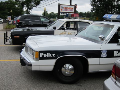 1991 Ford LTD Crown Victoria - Two white-and-black police cruisers park nose-out beneath a Dunkin' Donuts sign with an SUV behind them.