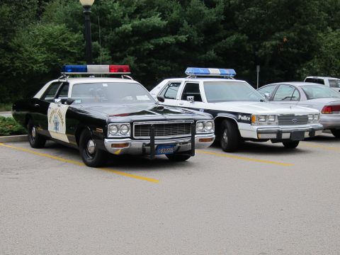 1991 Ford LTD Crown Victoria - black-and-white Highway Patrol sedan parks beside white police cruisers in a paved lot edged by trees.
