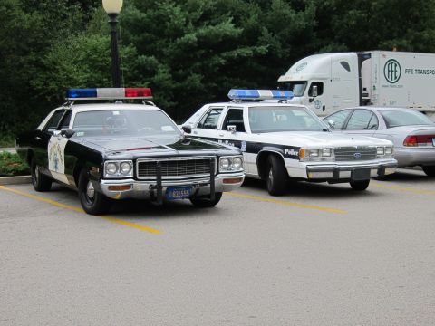 1991 Ford LTD Crown Victoria - black-and-white Highway Patrol sedan and a white police cruiser park in a lot with an FFE Transport semi behind.