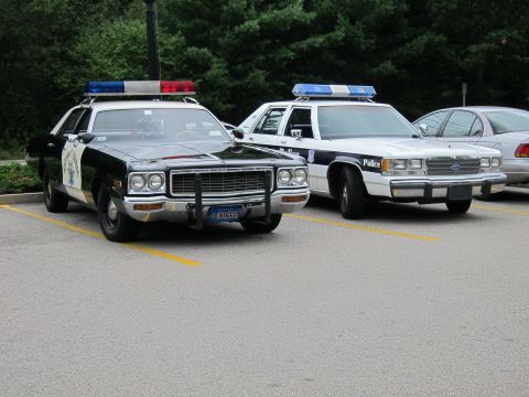 1991 Ford LTD Crown Victoria - black-and-white Highway Patrol sedan with chrome grille parks beside white police cruisers on striped pavement.