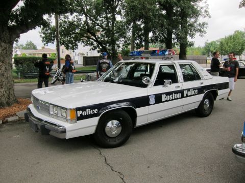 1991 Ford LTD Crown Victoria - white-and-black Ford four-door sedan lettered Boston Police parks under a tree with people and motorcycles behind.