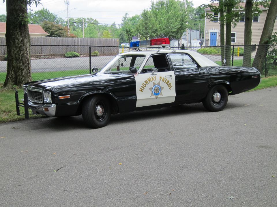 Black-and-white Highway Patrol sedan parked on a path by a chain-link fence