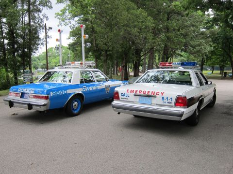 1991 Ford LTD Crown Victoria - blue NYPD sedan parks beside a white police cruiser lettered EMERGENCY CALL 9-1-1 on a tree-lined lot.