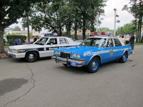 1991 Ford LTD Crown Victoria - blue NYPD police sedan with blue wheels and roof lights parks beside a white-and-black Boston Police cruiser.