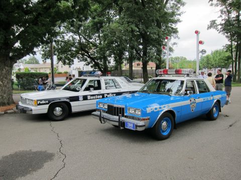 1991 Ford LTD Crown Victoria - blue NYPD sedan with a push bar parks beside a white-and-black Boston Police cruiser under shade trees.